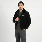 Front view of men's black velvet winter jacket with faux fur collar, model holding the lapel to show the fabric texture and gold chest logo.